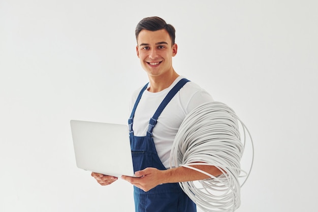 Utiliza portátil trabajador masculino en uniforme azul de pie dentro del estudio contra el fondo blanco.