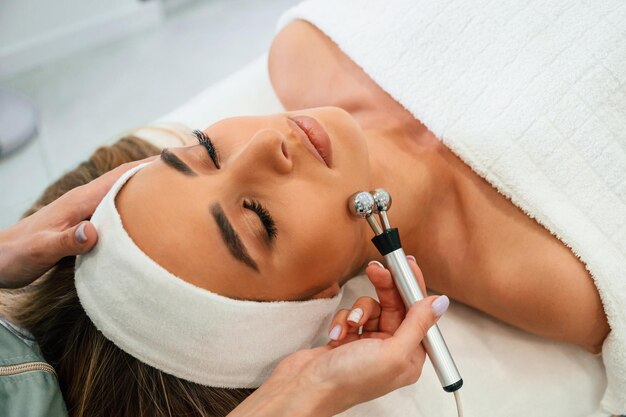 Se utiliza un rodillo de masaje para el rostro de una mujer que recibe cuidado facial por manos de una esteticista en un salón de spa