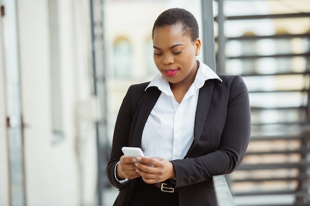 Utilizando smartphone empresaria afroamericana en traje de oficina sonriendo