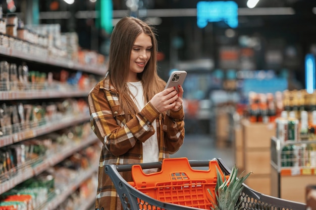 Utilizando el teléfono inteligente de pie La mujer está haciendo compras en el supermercado