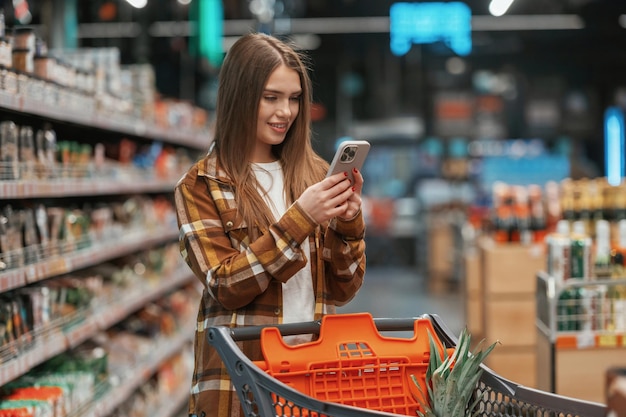 Foto utilizando el teléfono inteligente de pie la mujer está haciendo compras en el supermercado
