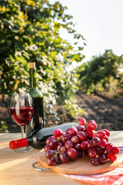 Uva fresca con botellas y copa de vino tinto sobre mesa de madera en viñedo