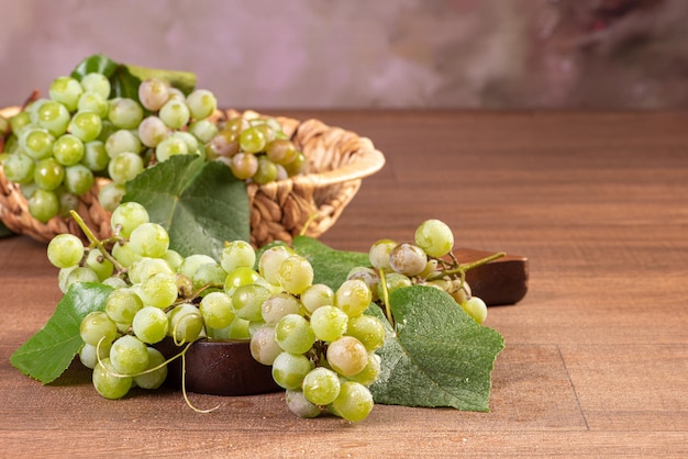 Uvas, cachos de uvas verdes colocados junto com uma cesta de palha na superfície de madeira, foco seletivo.