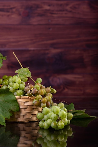 Uvas, cachos de uvas verdes colocados juntos com uma cesta de palha na superfície reflexiva, foco seletivo.