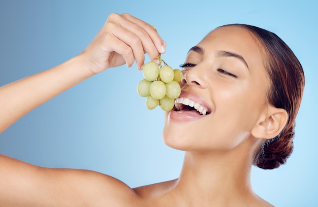 Uvas para a pele e mulher em estúdio para desintoxicação de bem-estar ou tratamento orgânico em fundo azul Lanche de frutas e modelo de menina com relaxamento durante a rotina de beleza e feliz com o produto cru isolado