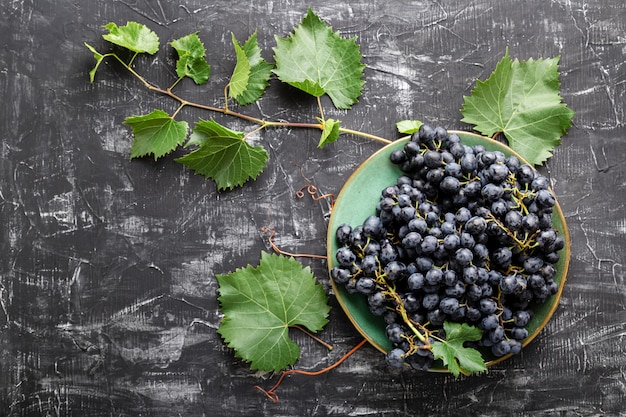 Uvas pretas no prato com a planta da videira na vista de cima da mesa escura. Comida doce de uvas suculentas no plano de fundo de concreto preto com espaço de cópia.