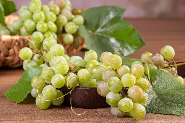 Uvas, racimos de uvas verdes colocados junto con una canasta de paja sobre la superficie de madera, enfoque selectivo.