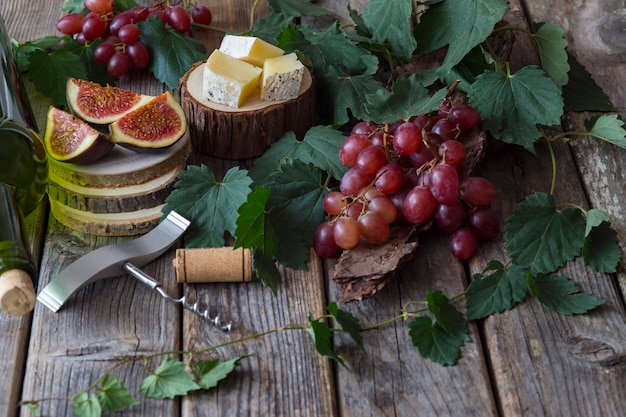 Uvas rojas, una botella de vino, un vaso, higos y queso en un soporte de madera, una botella de vino