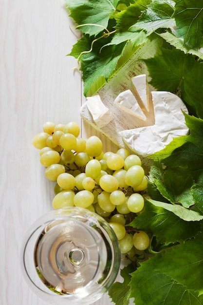 Uvas verdes, queijo camembert fresco e copo de vinho branco sobre o fundo branco de madeira