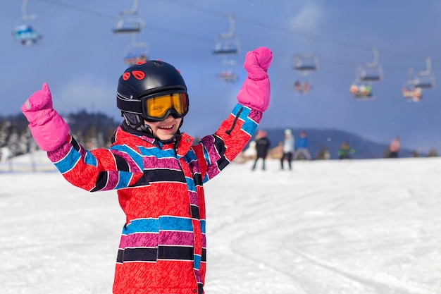 Vacaciones de esquí para niños en las montañas de invierno. Niña feliz en traje de esquí Gafas Casco cerca del remonte