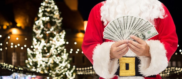 Foto vacaciones, finanzas y concepto de personas - primer plano de papá noel con dinero en dólares sobre el fondo del árbol de navidad