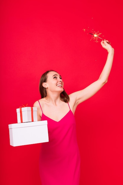 Vacaciones de humor de Año Nuevo Retrato de una mujer joven en un vestido de noche rosa con bengalas y sosteniendo regalos en cajas sobre un fondo rojo en el estudio