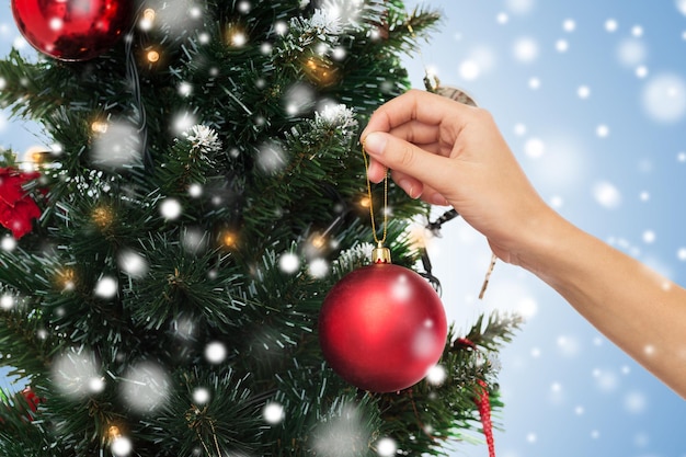 vacaciones de invierno, celebración y concepto de la gente - cierre de la mano decorando el árbol de navidad con bola sobre fondo azul con nieve