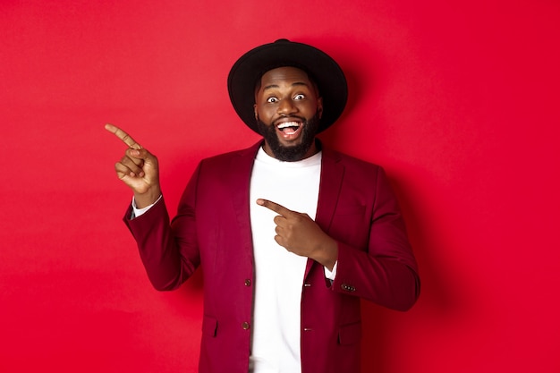 Vacaciones de invierno y concepto de compras. Feliz el hombre negro apuntando a la izquierda y sonriendo, mostrando la oferta de promoción de año nuevo sobre fondo rojo.