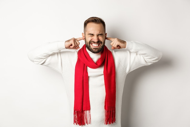 Vacaciones de invierno y concepto de compras. Hombre molesto cerró los oídos con los dedos, angustiado por ruidos fuertes o vecinos, de pie contra el fondo blanco.