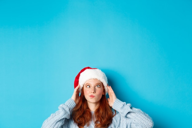 Vacaciones de invierno y concepto de Nochebuena. Cabeza de niña pelirroja divertida con sombrero de santa, aparece desde abajo y entrecerrando los ojos, haciendo muecas, de pie cerca del espacio de la copia sobre fondo azul.