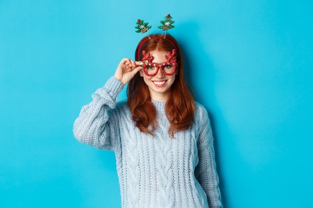 Vacaciones de invierno y concepto de ventas de Navidad. Modelo de mujer hermosa pelirroja celebrando el año nuevo, con gafas y diadema de fiesta divertida, sonriendo a la cámara.
