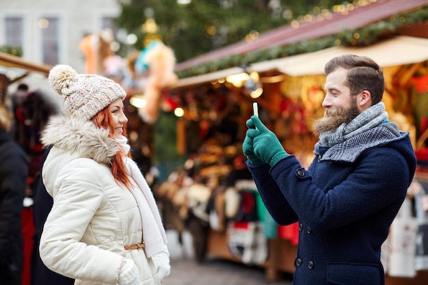 vacaciones, invierno, navidad, tecnología y concepto de personas - feliz pareja de turistas vestidos de abrigo tomando selfie con smartphone en el casco antiguo