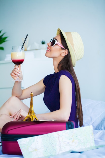 Vacaciones. Mujer que se prepara para descansar. La muchacha hermosa joven se sienta en la cama y se sostiene en el cóctel de las manos. Retrato de una mujer sonriente. Niña feliz se va de vacaciones