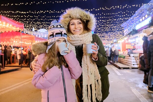 Vacaciones de Navidad y año nuevo, feliz madre e hija niño caminando juntos bebiendo té caliente en el mercado de Navidad, luces brillantes de guirnaldas de la noria de la ciudad por la noche