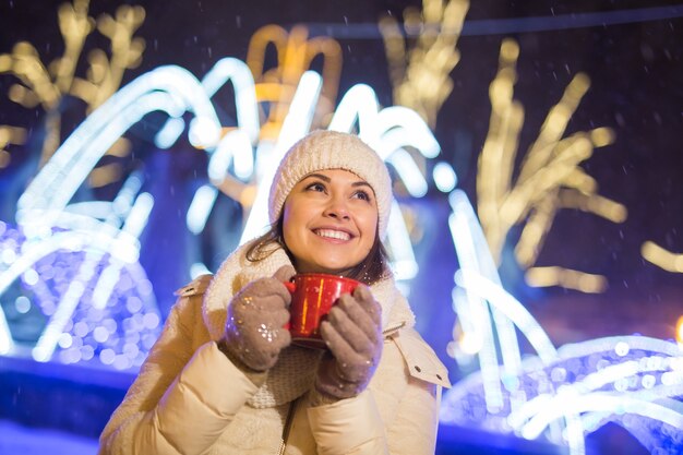Vacaciones de Navidad hermosa mujer sonriente en ropa de abrigo con una taza de café caliente al aire libre en invierno