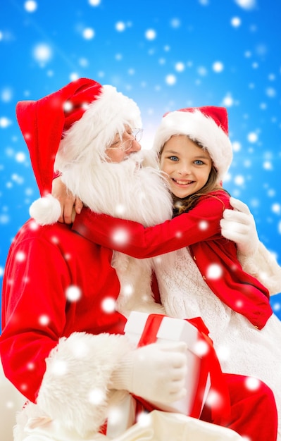 vacaciones, navidad, infancia y concepto de la gente - niña sonriente abrazándose con santa claus sobre fondo azul nevado