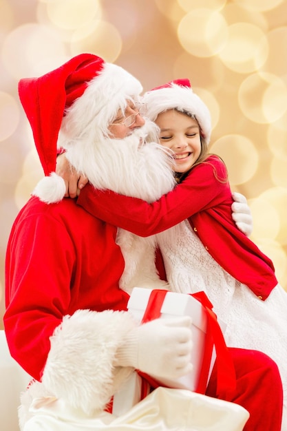 vacaciones, navidad, infancia y concepto de la gente - niña sonriente abrazándose con santa claus sobre fondo de luces beige