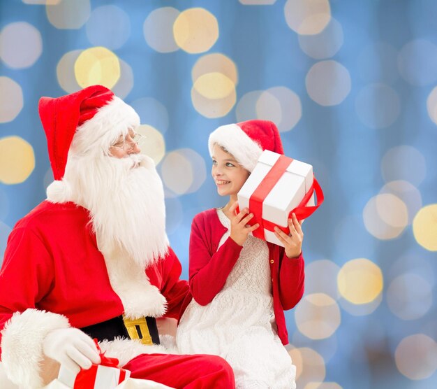 vacaciones, navidad, infancia y concepto de la gente - niña sonriente con santa claus y regalos sobre fondo de luces azules