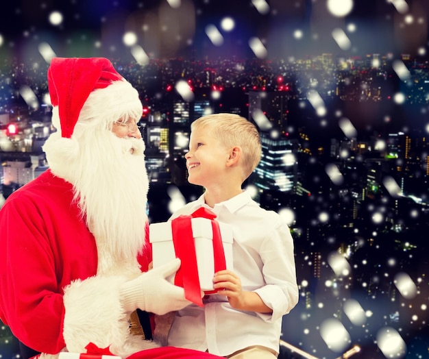 vacaciones, navidad, infancia y concepto de la gente - niño sonriente con santa claus y regalos sobre el fondo de la ciudad nevada