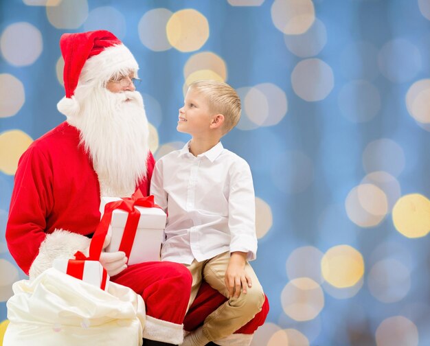 vacaciones, navidad, infancia y concepto de la gente - niño sonriente con santa claus y regalos sobre fondo de luces azules