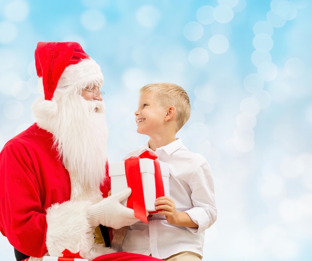 vacaciones, navidad, infancia y concepto de la gente - niño sonriente con santa claus y regalos sobre fondo de luces azules