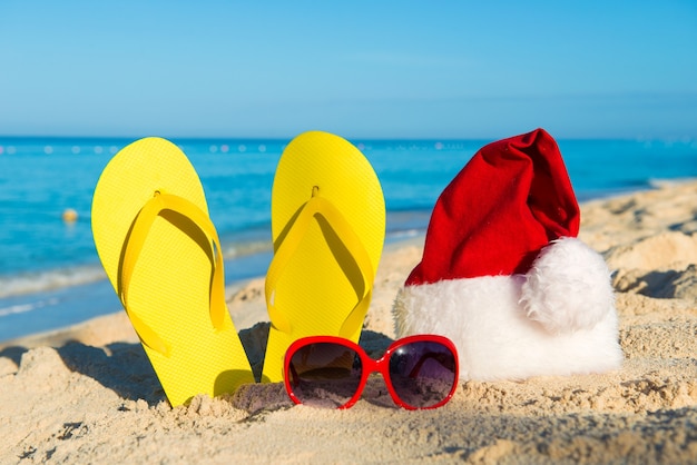 Vacaciones de Navidad en el mar. Feliz año nuevo. Gorro de Papá Noel, sandalias, gafas de sol en la playa de arena