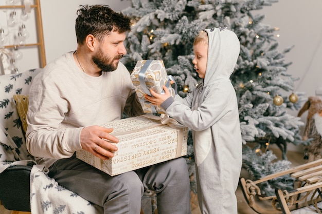 Vacaciones de Navidad Padre guapo jugando con un pequeño hijo lindo cerca del árbol de año nuevo decorado en casa Niño de tradición familiar le da un regalo a su padre