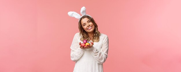 Vacaciones primavera y concepto de fiesta Retrato tierna y romántica chica rubia con vestido blanco y orejas de conejo inclinan la cabeza linda sonriendo feliz como sosteniendo huevos pintados celebrando el fondo rosa de Pascua