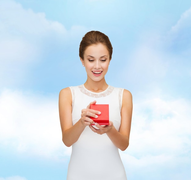 vacaciones, regalos, boda y concepto de felicidad - mujer sonriente con vestido blanco sosteniendo una caja de regalo roja sobre fondo azul cielo nublado