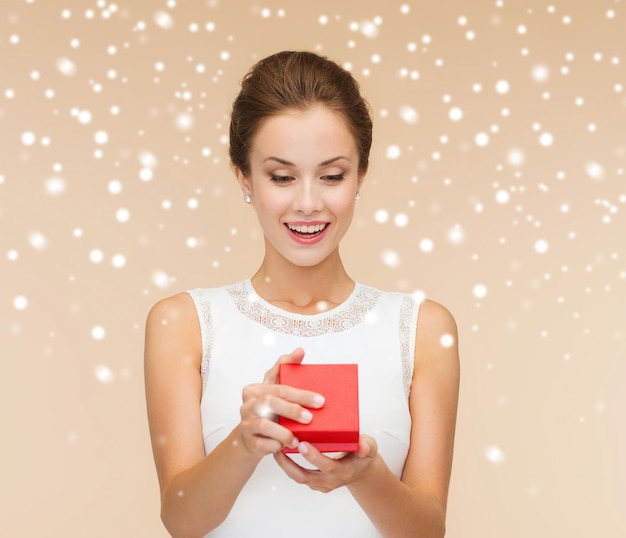 vacaciones, regalos, boda y concepto de felicidad - mujer sonriente con vestido blanco sosteniendo una caja de regalo roja sobre fondo beige sobre fondo beige y nieve