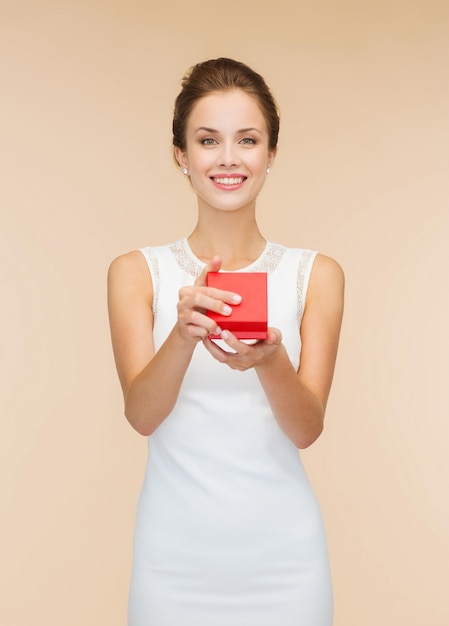 vacaciones, regalos, boda y concepto de felicidad - mujer sonriente con vestido blanco sosteniendo una caja de regalo roja sobre fondo beige