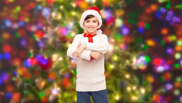 vacaciones, regalos, navidad, infancia y concepto de la gente - niño feliz sonriente con sombrero de santa con caja de regalo durante las vacaciones luces de fondo