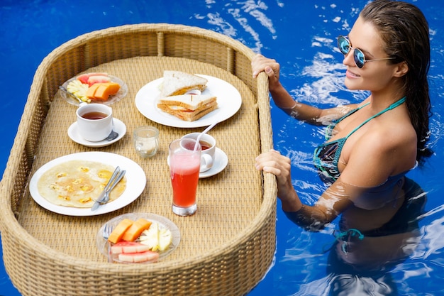 Vacaciones en el resort. Mujer joven feliz con un desayuno flotante en la piscina.