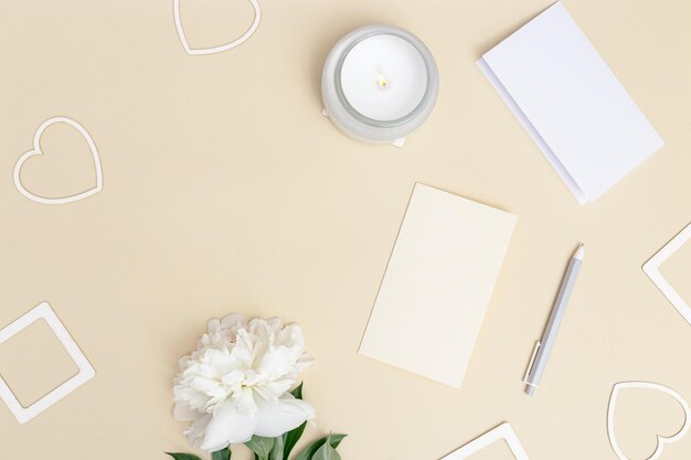 Vacaciones románticas o día de San Valentín con flores de peonía frescas, tarjetas para deseos de celebración, bolígrafo, corazones, velas aromáticas encendidas sobre fondo beige. Amor, concepto romántico.