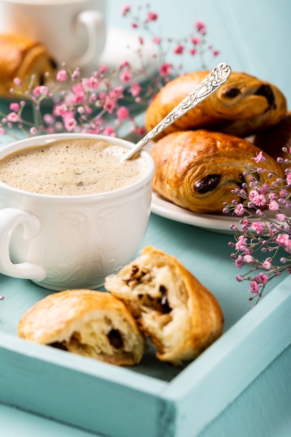 Vacaciones con una taza de café, mini croissants recién hechos, bollo de chocolate y flores de clavel sobre una superficie turquesa