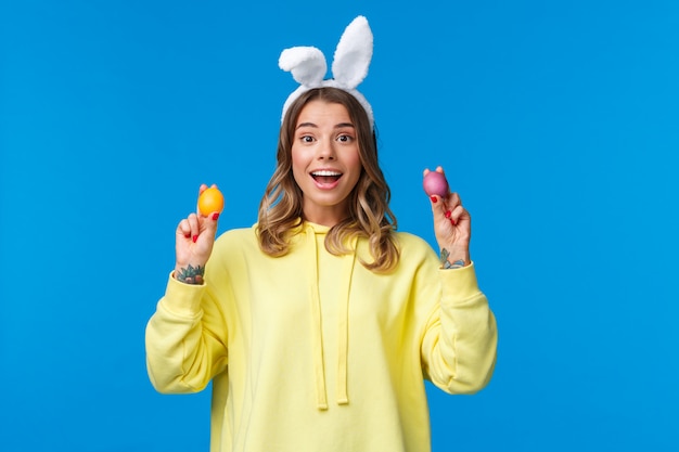 Vacaciones, tradiciones y concepto de celebración. Linda rubia caucásica hermana quiere jugar con hermano sosteniendo dos huevos de Pascua pintados y sonriente, con orejas de conejo, pared azul