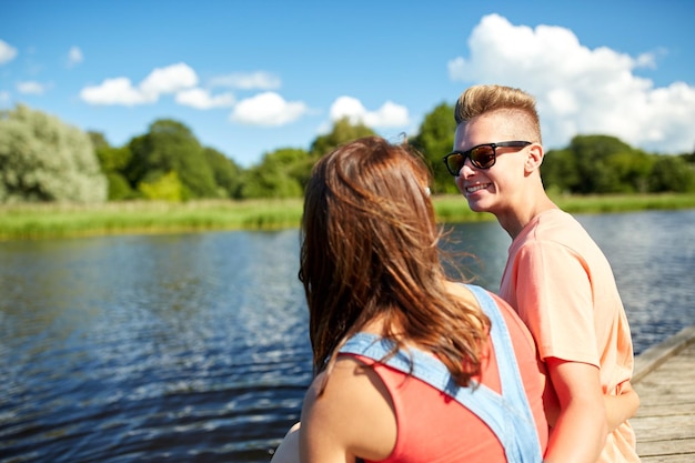 vacaciones, vacaciones, amor y concepto de la gente - feliz pareja adolescente sentada en el muelle del río en verano