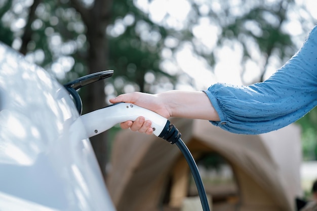 Foto vacaciones de vacaciones de camping viajando a la naturaleza con coche eléctrico mujer recargar coche ev desde la estación de carga en el campamento viajes naturales y coche ecológico para un medio ambiente sostenible perpetuo.