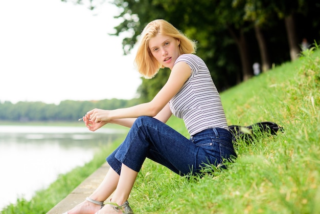 Vacaciones de verano. Chica relajándose en la orilla del río después de la jornada laboral. Mujer soñando con vacaciones. Descansa, relájate y hobby. Dedique tiempo libre para usted. La muchacha se sienta en la hierba verde cerca del río. Temporada de vacaciones.