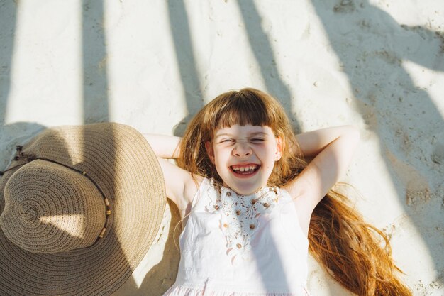 Vacaciones de verano y concepto de la gente niña en un sombrero de paja tomando el sol en una playa de arena