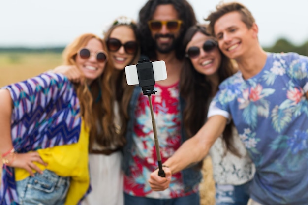 vacaciones de verano, tecnología y gente concepto - sonrientes jóvenes amigos hippie con gafas de sol tomando una foto con un teléfono inteligente en un selfie stick en el campo