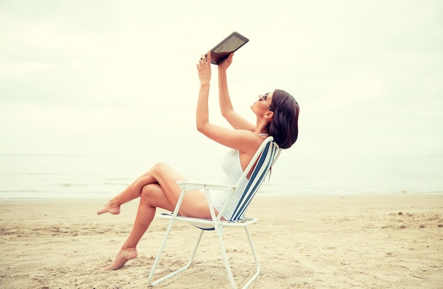 vacaciones de verano, turismo, viajes, vacaciones y concepto de personas - mujer joven sonriente con computadora de tablet pc sentada en una silla plegable y tomando selfie en la playa