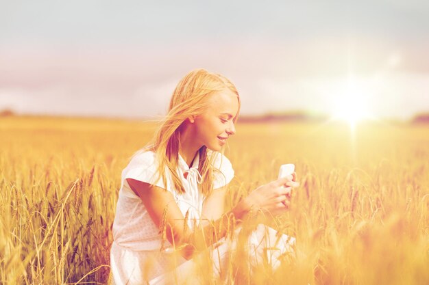 vacaciones de verano, vacaciones, tecnología y concepto de personas - joven sonriente vestida de blanco con smartphone y auriculares escuchando música en el campo de cereales