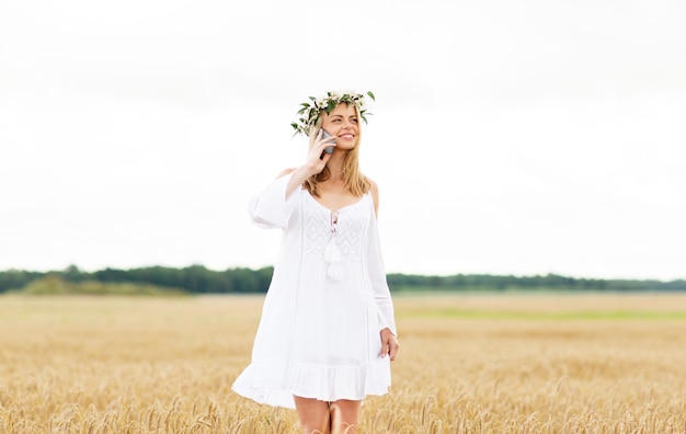 vacaciones de verano, vacaciones, tecnología y concepto de personas - mujer joven sonriente en corona de flores llamando al teléfono inteligente en el campo de cereal